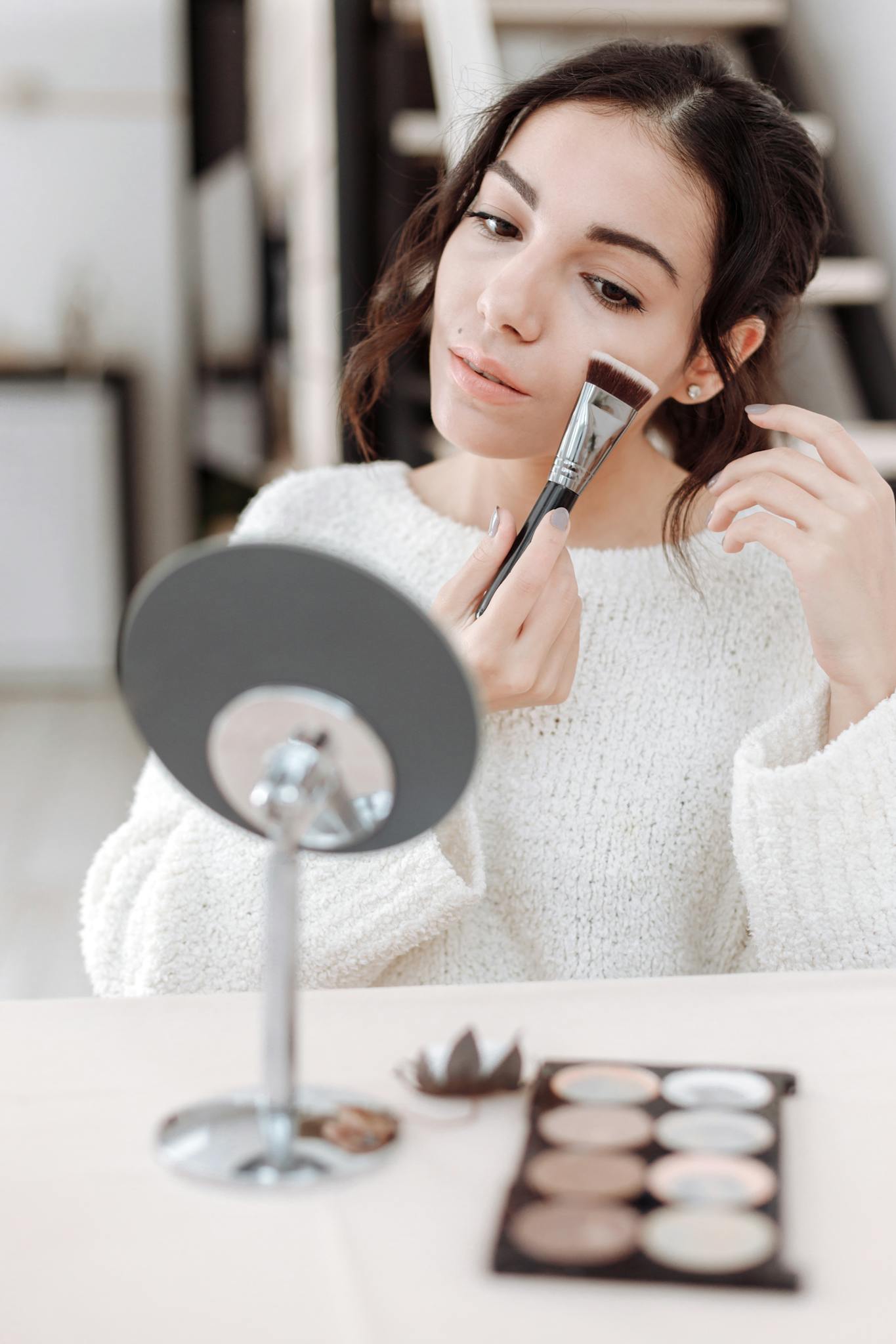 Woman using makeup brush with mirror and palette, creating a beauty routine.